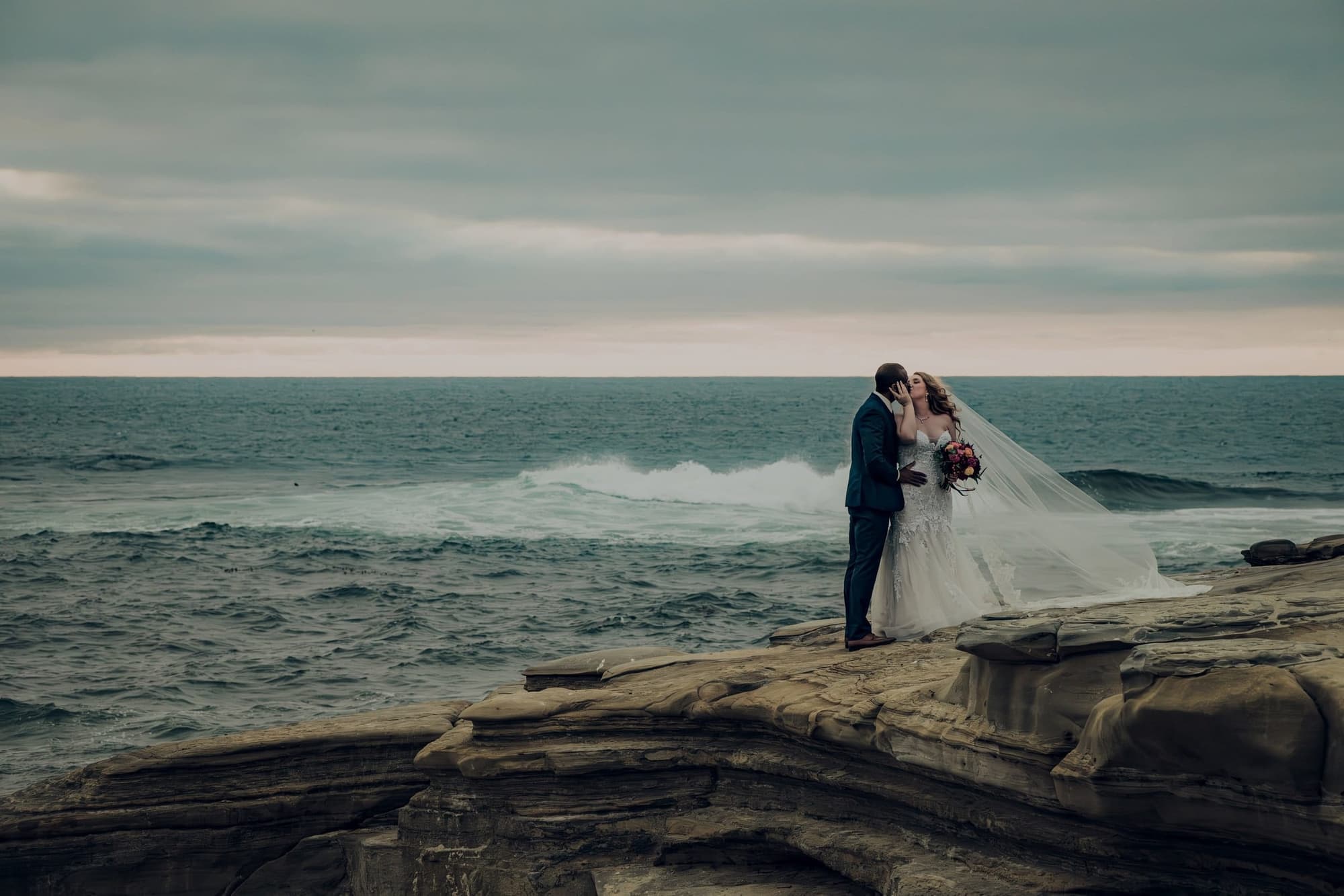 Bride and groom kissing on a coastal cliff at dusk